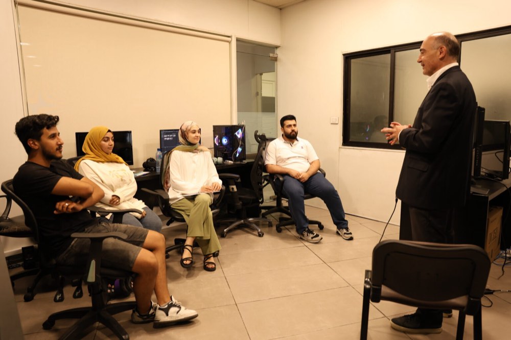 Fadi Tuffaha, in a dark suit, standing and speaking with his hands open to four participants seated on office chairs in a small training room lined with computer workstations.